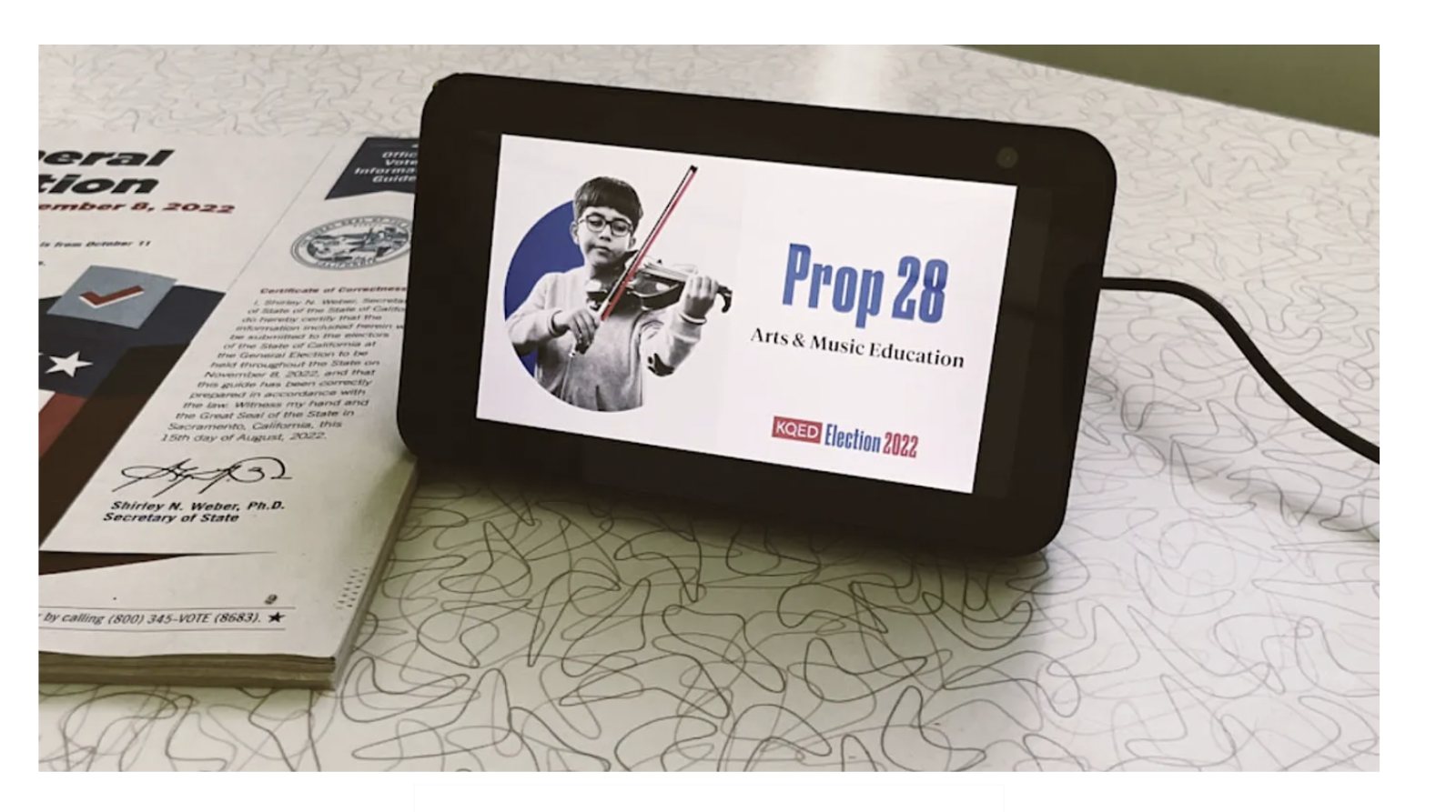 An Echo Show displaying a KQED Election 2022 card for Prop 28, Arts and Music Education, resting beside the official California voter information guide